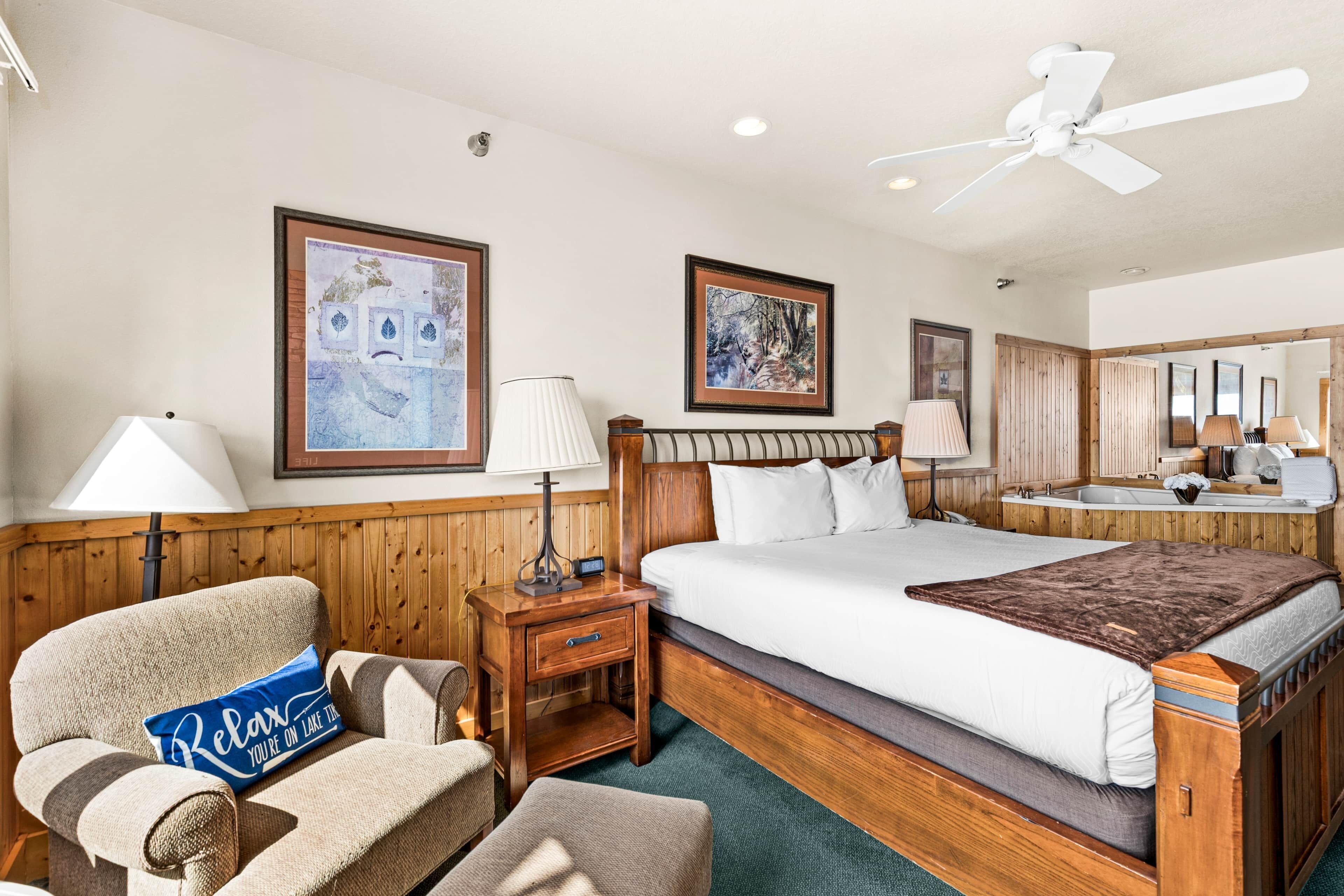 A hotel room with a large bed, a wooden headboard, a nightstand with a lamp, a chair with a pillow, and framed artwork on the walls.