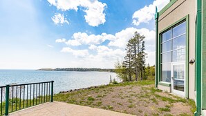 A paved patio with a view of a lake and distant land, a green building with a glass door, and a metal railing.