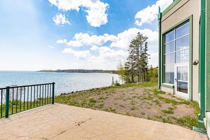 A paved patio with a view of a lake and distant land, a green building with a glass door, and a metal railing. - Majestic Lake Views | 1BR w/King Suite | Pools (Two Harbors)