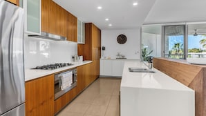 A modern kitchen with wooden cabinets, a stainless steel refrigerator, a built-in oven, a gas stove, and a large window offering a view of palm trees.