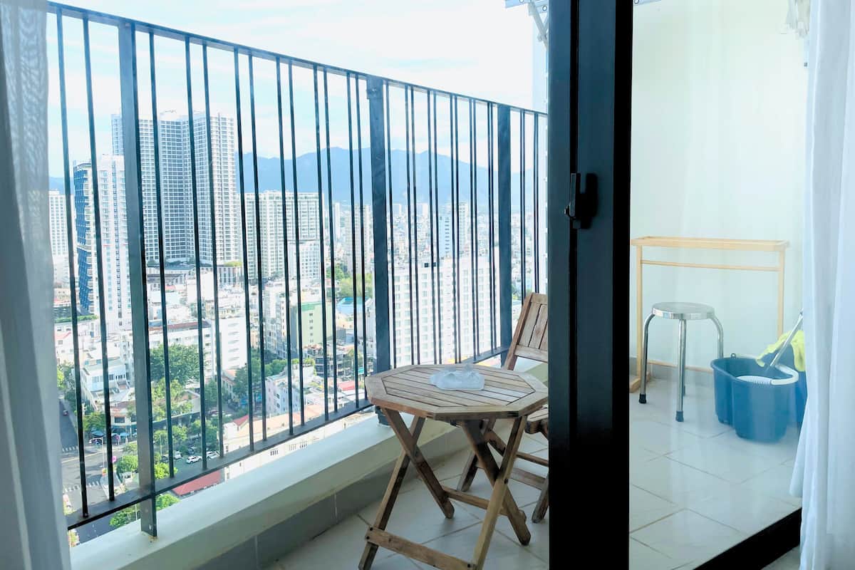Un balcón con vistas a un paisaje urbano, una silla de madera, una mesita pequeña y una cesta de ropa azul.