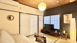 Un salón de estilo japonés tradicional con suelo de tatami, una mesa de madera baja, un sofá, un televisor y un panel shoji.