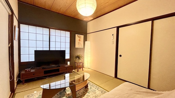 Una habitación japonesa tradicional con suelo de tatami, techo bajo de madera, un televisor sobre un soporte de madera, una mesa de centro de vidrio y puertas correderas.