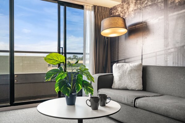 A modern living room with a grey sofa, a round table with a potted plant and two mugs, and a large window offering a view of the sky and buildings.