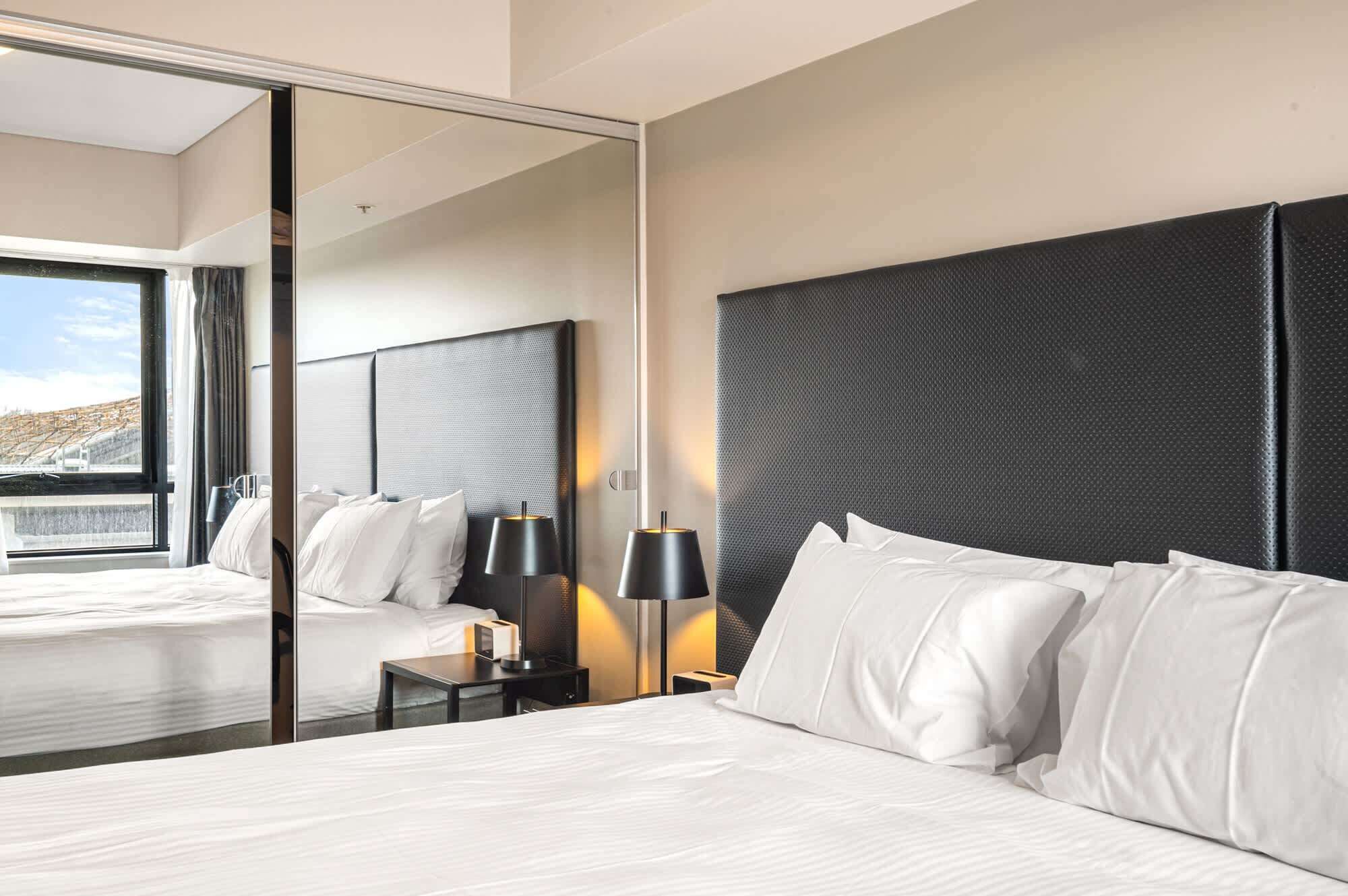 Ein Hotelzimmer mit einem großen Bett, einem Nachttisch mit Lampe, einem Spiegel und Blick auf eine Stadtlandschaft.