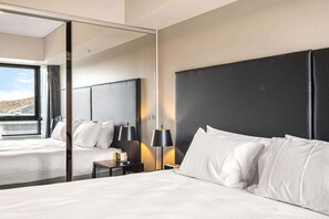 A hotel room with a large bed, a nightstand with a lamp, a mirror, and a view of a cityscape.