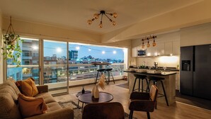 A modern apartment with a living room, dining area, and kitchen. It features large windows with a city view, a hanging plant, and a unique chandelier. - Charming 3-bedroom apartment with rooftop pool in Barranco (Barranco)