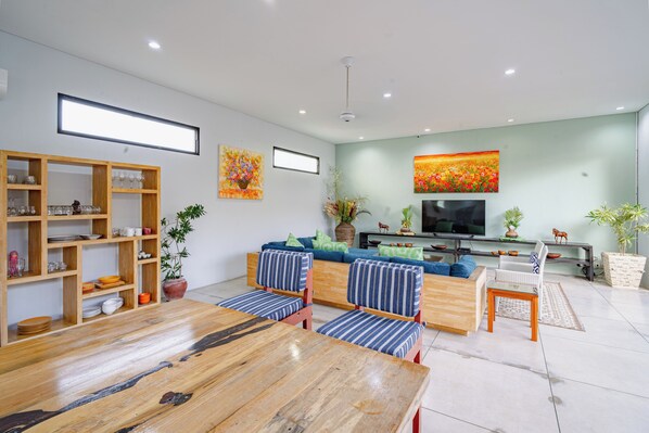 A modern living room with a wooden dining table, sofas, a TV, and a bookshelf.