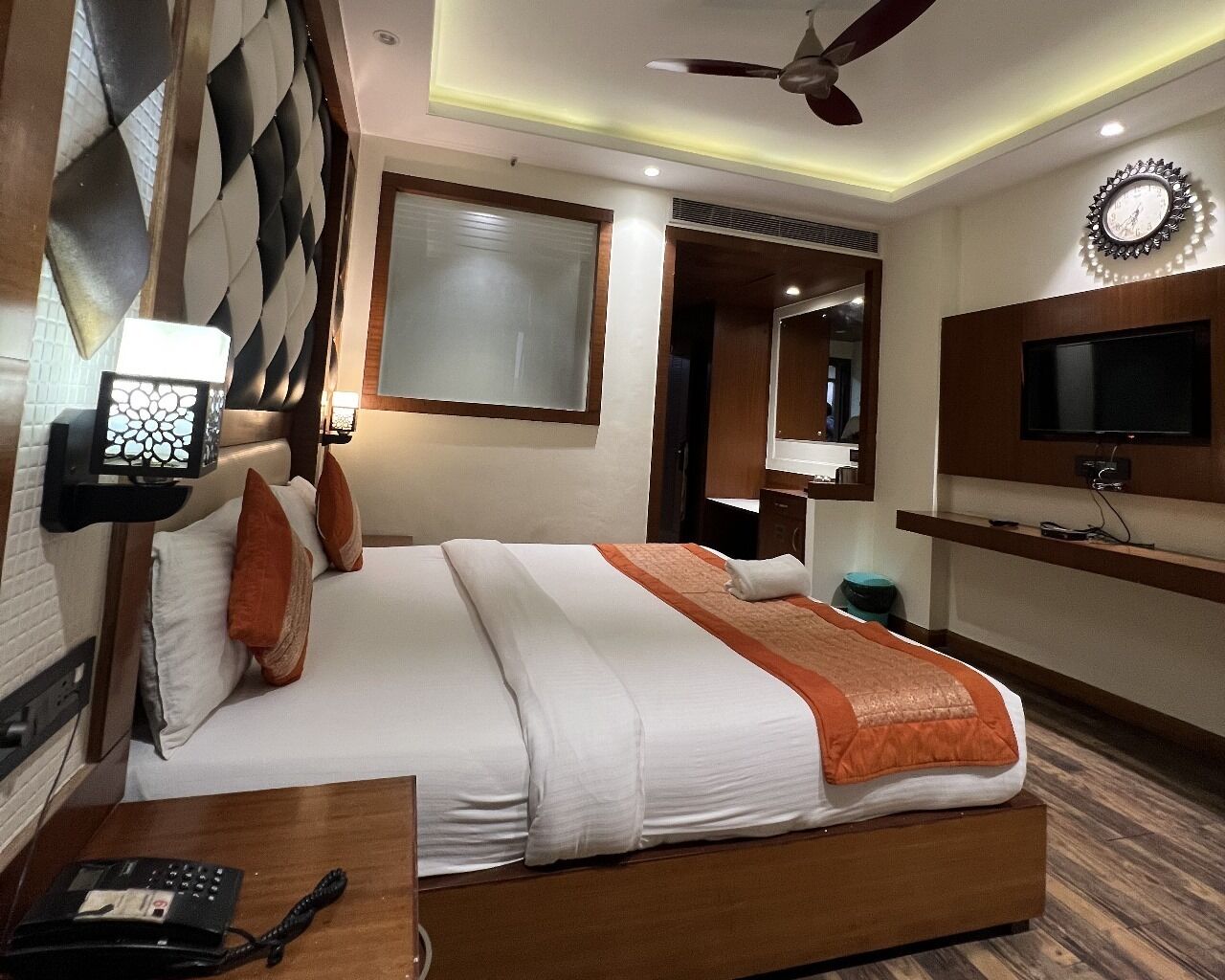 Una habitación de hotel con cama, televisión, reloj y ventilador de techo.