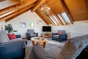 Una habitación con techos altos y estructura de vigas de madera, televisor y disposición de asientos que incluye sofás y una silla.