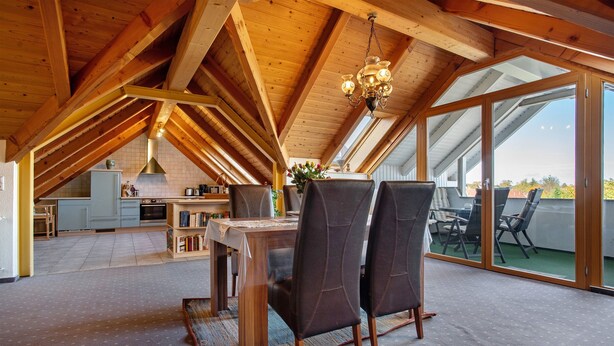 En rummelig spiseafdeling med højt til loftet og træbjælker, et køkken i baggrunden og store skydedøre til en udendørs terrasse.