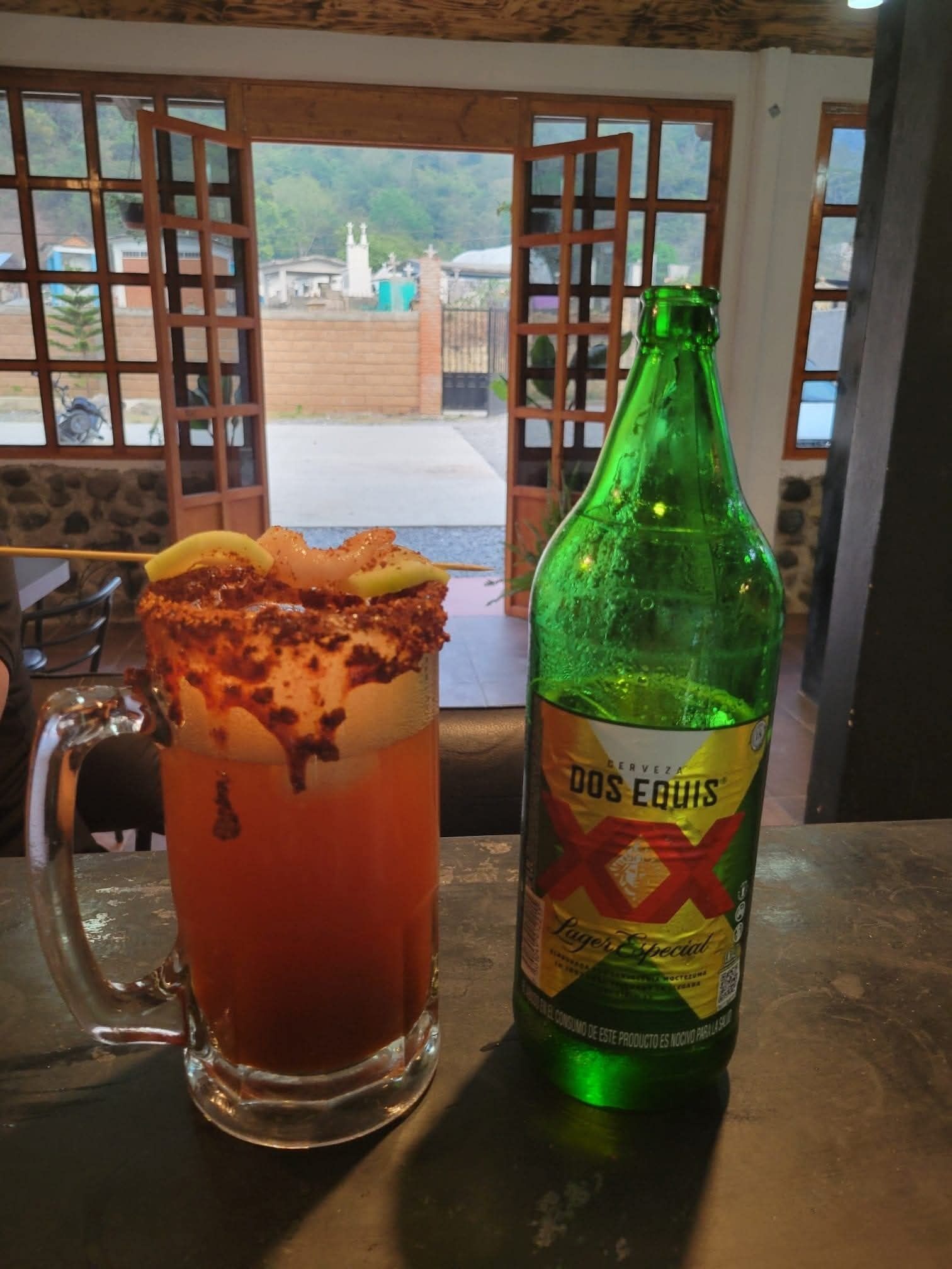 Un vaso de té helado con una rodaja de limón y una rama de canela, junto a una botella de cerveza Dos Equis sobre una mesa con vista a un edificio a través de una ventana.