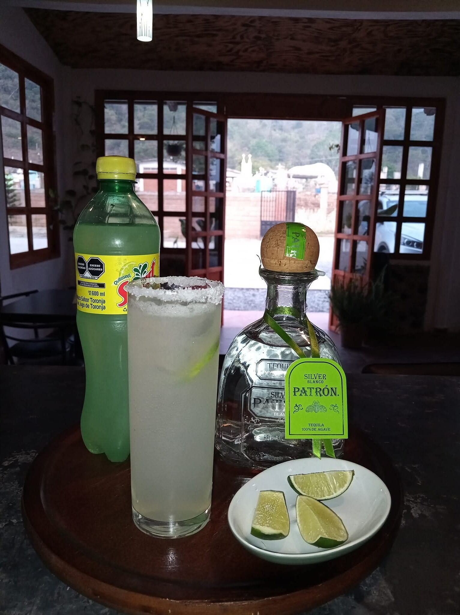 Una mesa con una botella de jugo de Toronja, un vaso de un líquido transparente, una botella de tequila Silver Patron y un plato con rebanadas de lima.