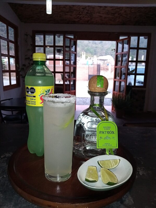 Une table avec une bouteille de jus de pamplemousse, un verre contenant un liquide transparent, une bouteille de tequila Silver Patron et une assiette avec des tranches de citron vert.