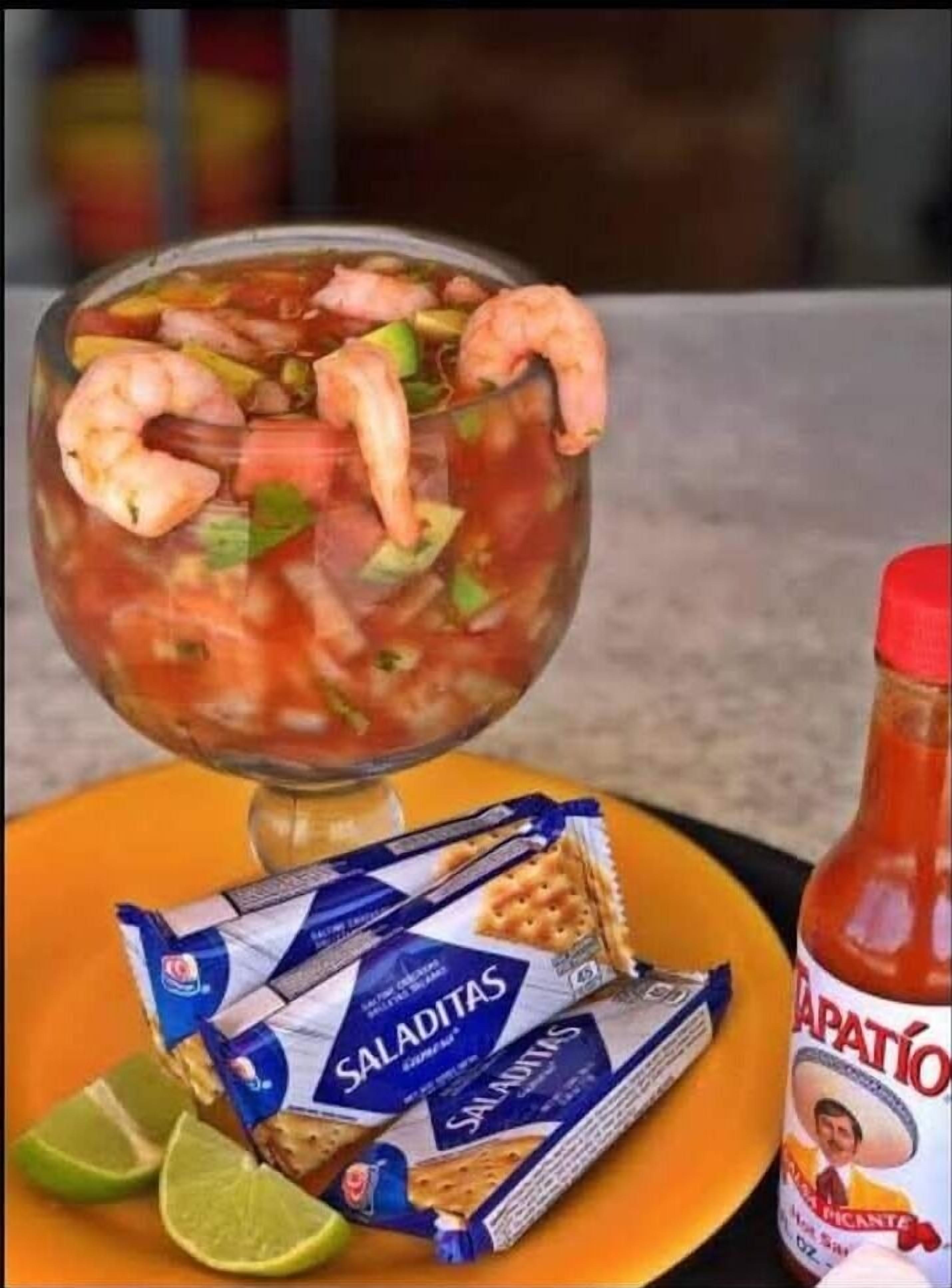 A shrimp cocktail in a glass, accompanied by a packet of 'Saladitas' crackers, a bottle of 'Dapatio' sauce, and lime wedges on a table.