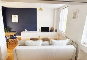Ett modernt vardagsrum med en vit soffa, en matplats med bord och stolar, en tv och en mörk accentvägg med en inramad tavla.