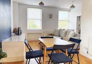 A modern living room with a dining table, chairs, a sofa, and two windows with blinds.