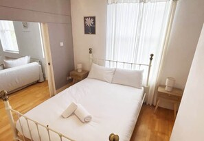 Un dormitorio con cama, mesita de noche, lámpara de pared y ventana con cortinas.