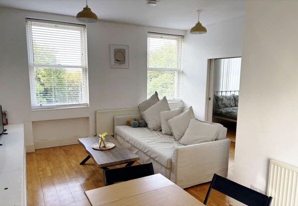 A living room with a sofa, a dining table, and two windows with blinds.