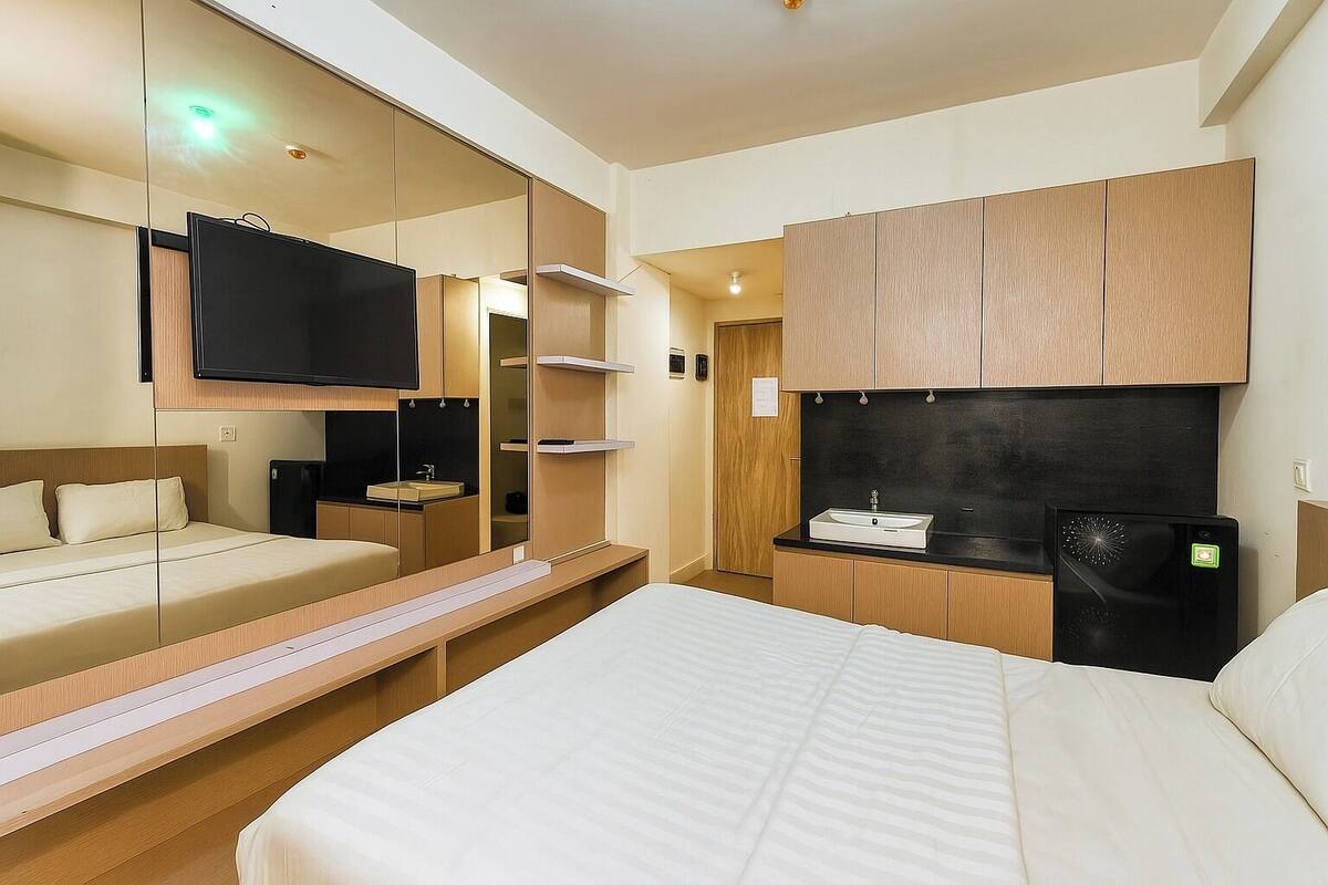 A modern hotel room with a large bed, a TV mounted on a mirrored wall, a small kitchenette, and a walk-in closet.