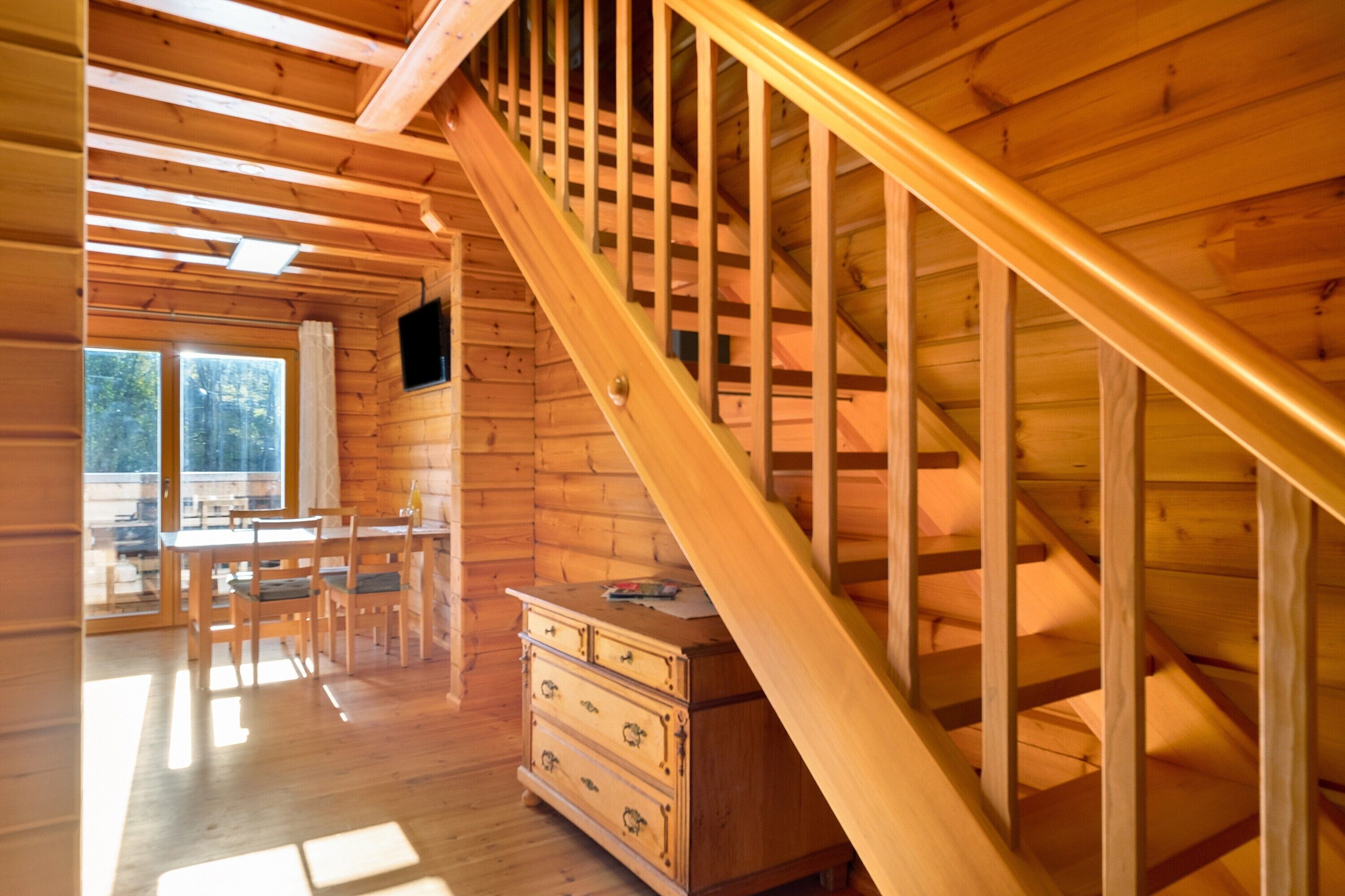 A wooden interior with a staircase, a dining area with chairs and a table, and a dresser.