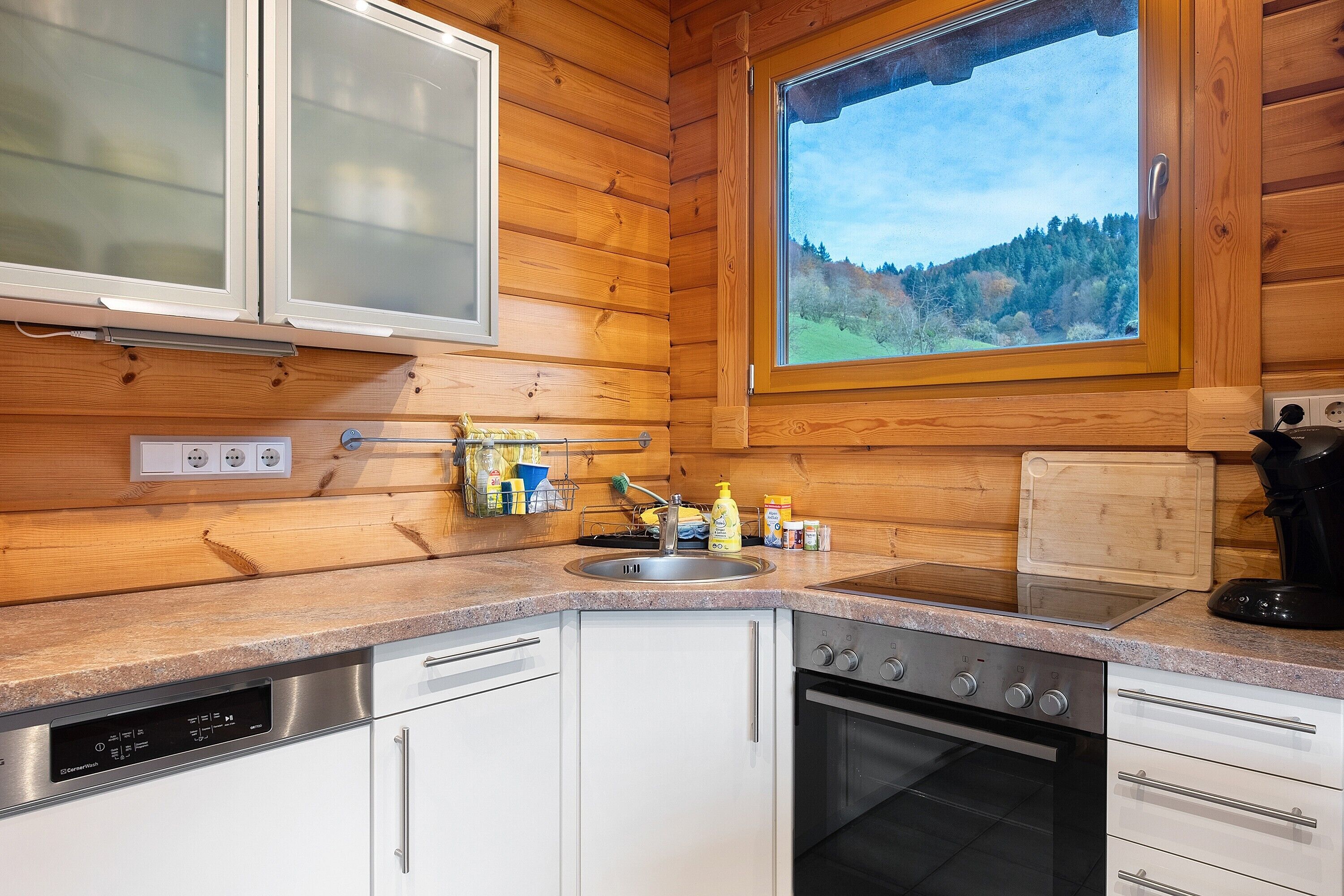 A kitchen with wooden walls, a window with a view, a sink, a dishwasher, and an oven.