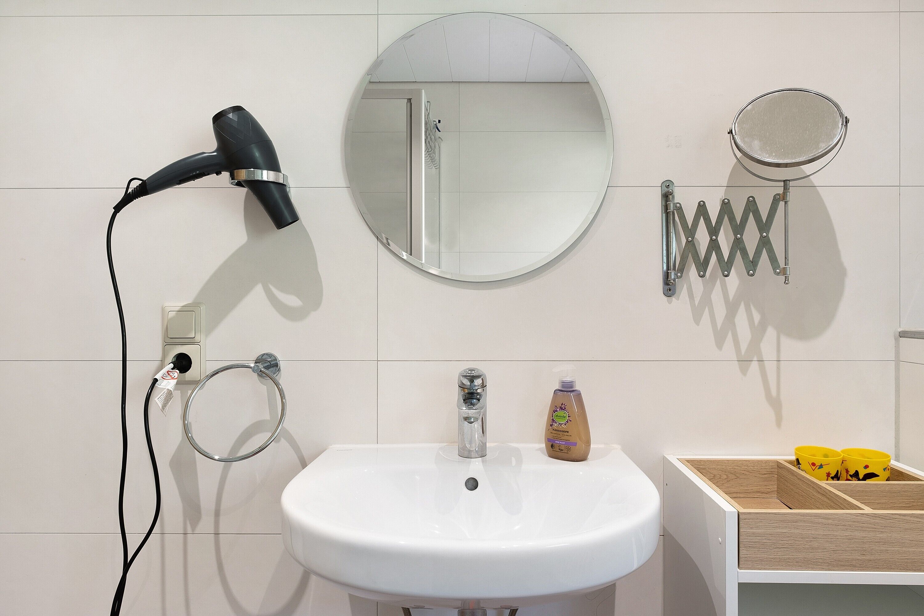 Un baño con lavamanos blanco, secador de pelo, espejo, dispensador de jabón y un mueble de almacenamiento de madera.