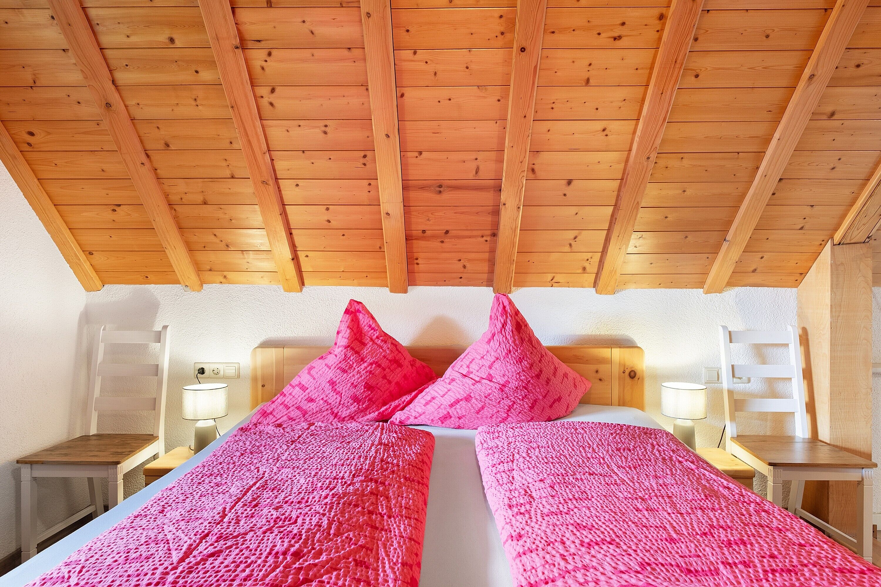 Un plafond en bois, un lit avec une literie rose, deux tables de chevet et deux chaises.