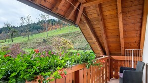 A wooden balcony with a view of a green hillside, a wicker chair, and a wooden railing. - Apartment 'Talblick' with Shared Terrace, Garden, and Wi-Fi (Gengenbach)