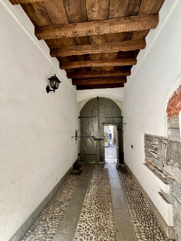 Un corridoio con pavimento in pietra, travi di legno al soffitto e un'ampia porta ad arco.