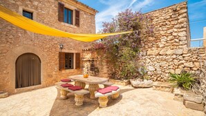 Un patio de piedra con una mesa redonda y bancos, rodeado de muros de piedra y un toldo amarillo.