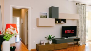 A modern living room with a wooden floor, a TV displaying 'NETFLIX,' a bookshelf, a plant, and a vase with flowers.