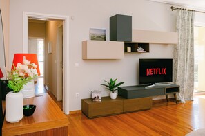 A modern living room with a wooden floor, a TV displaying 'NETFLIX,' a bookshelf, a plant, and a vase with flowers.