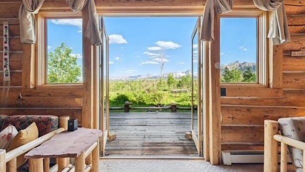 Una baita di legno con un terrazzo in legno che conduce a una grande finestra, con vista su un paesaggio montuoso.