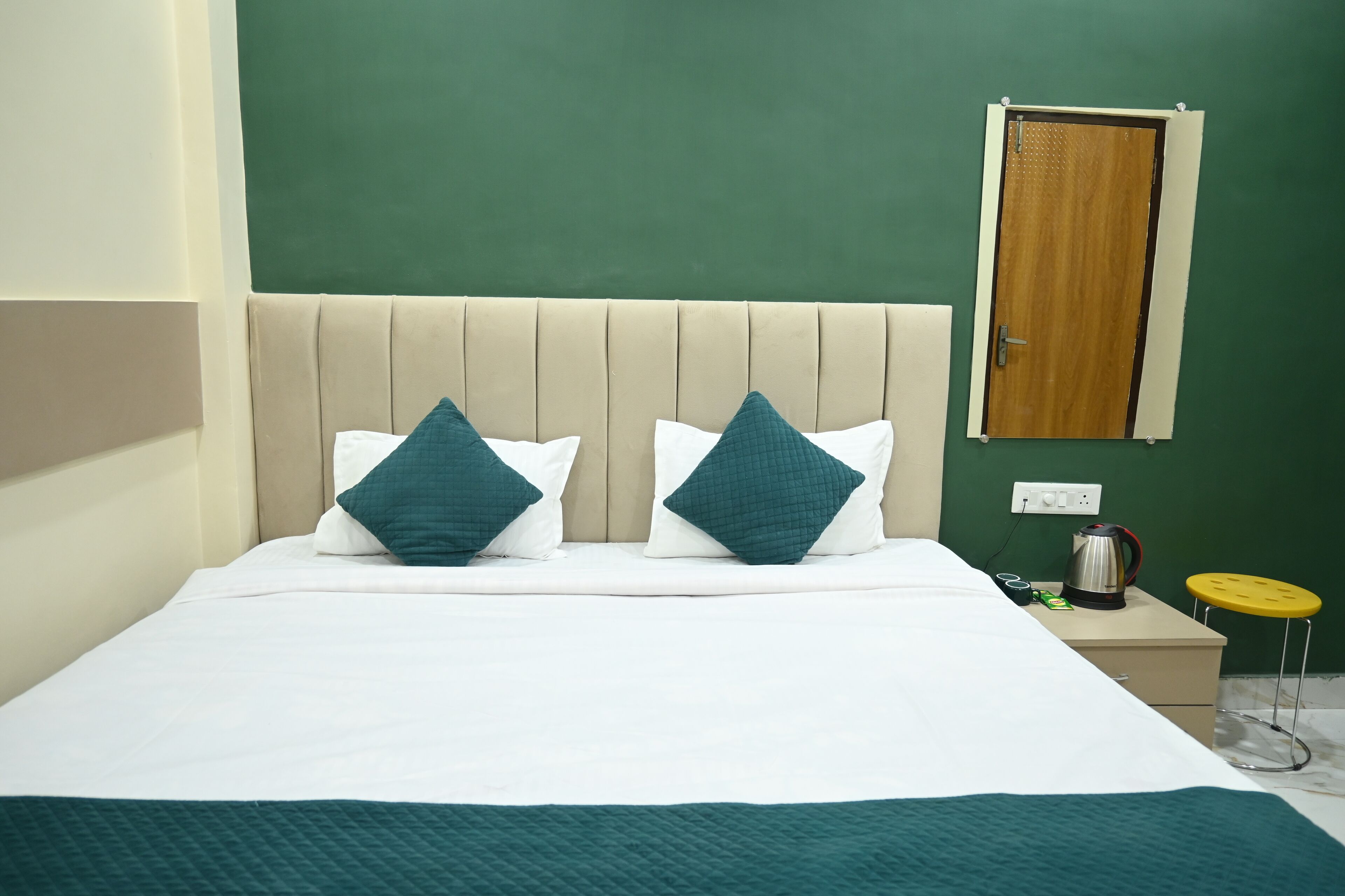 A neatly made bed with white and teal bedding, a beige headboard, a small bedside table with a kettle and a yellow stool, and a green wall with a wooden door.