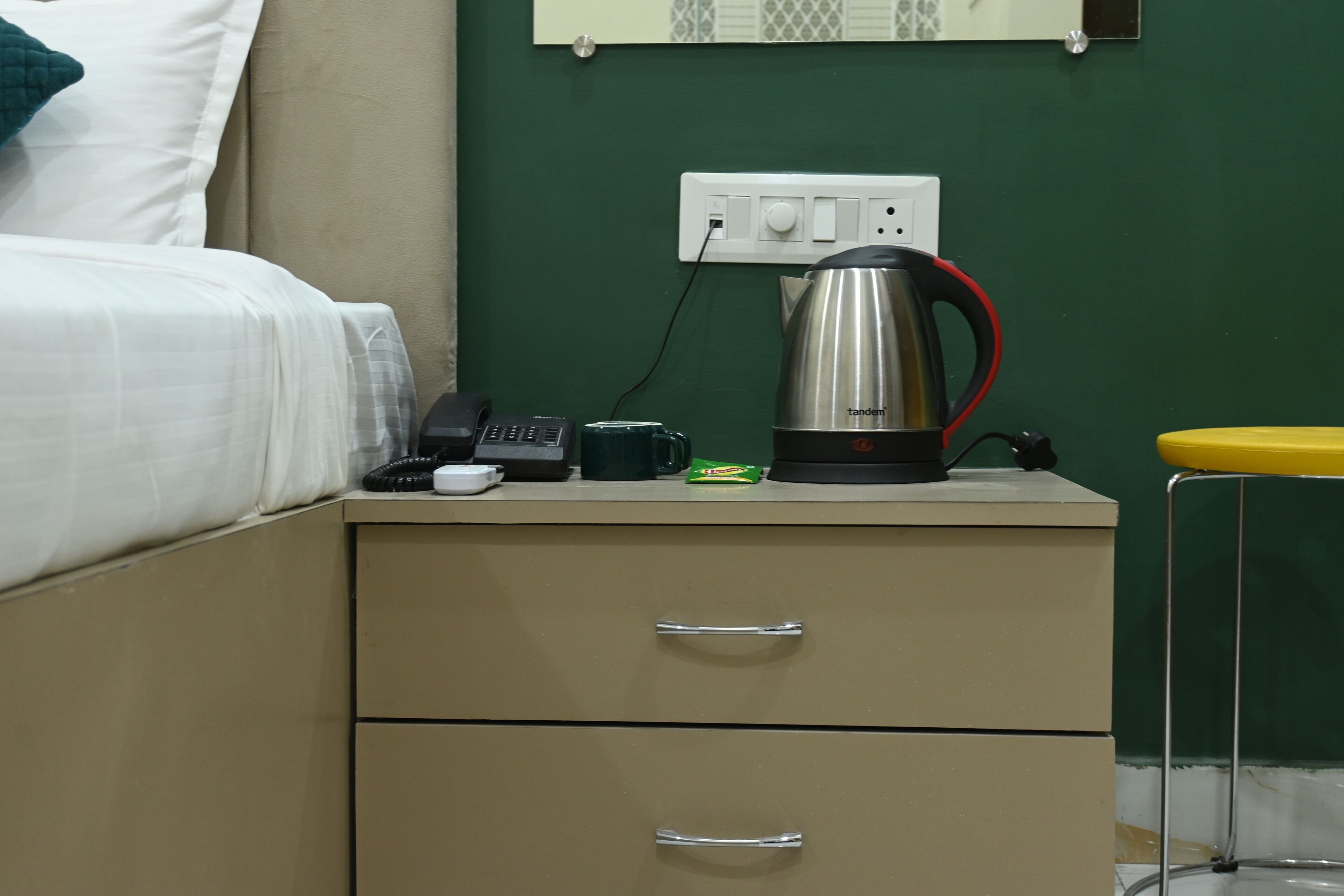 A hotel room with a bed, a bedside table with a kettle, a phone, and a cup.