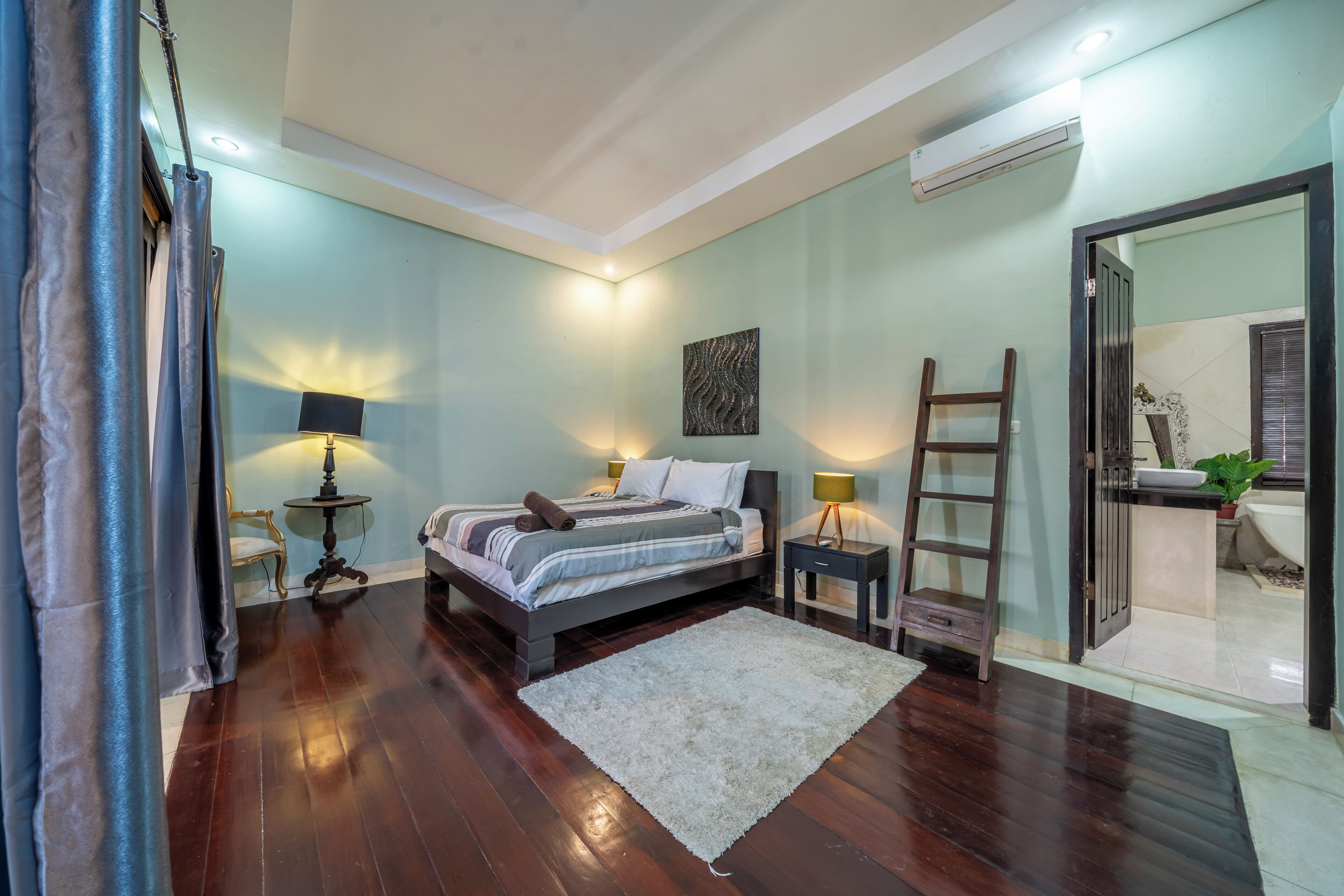 A bedroom with a bed, bedside tables, a nightstand, a ladder, and a bathroom visible through an open door.