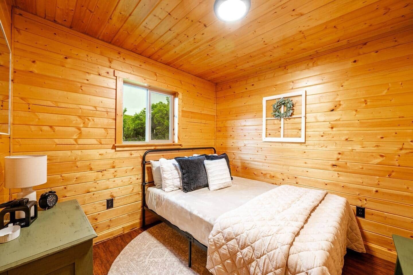 Quarto com painéis de madeira, uma cama, uma janela, um arranjo fixado na parede e uma mesa de cabeceira com abajur e relógio.
