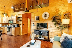 A wooden interior with a kitchen, dining area, and living room, featuring a clock, plants, and a fireplace.