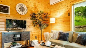 Gemütliches Interieur einer Holzhütte mit Holzwand, einer Uhr, einem Fernseher, einer Couch und einem Fenster mit Blick auf Grünflächen.