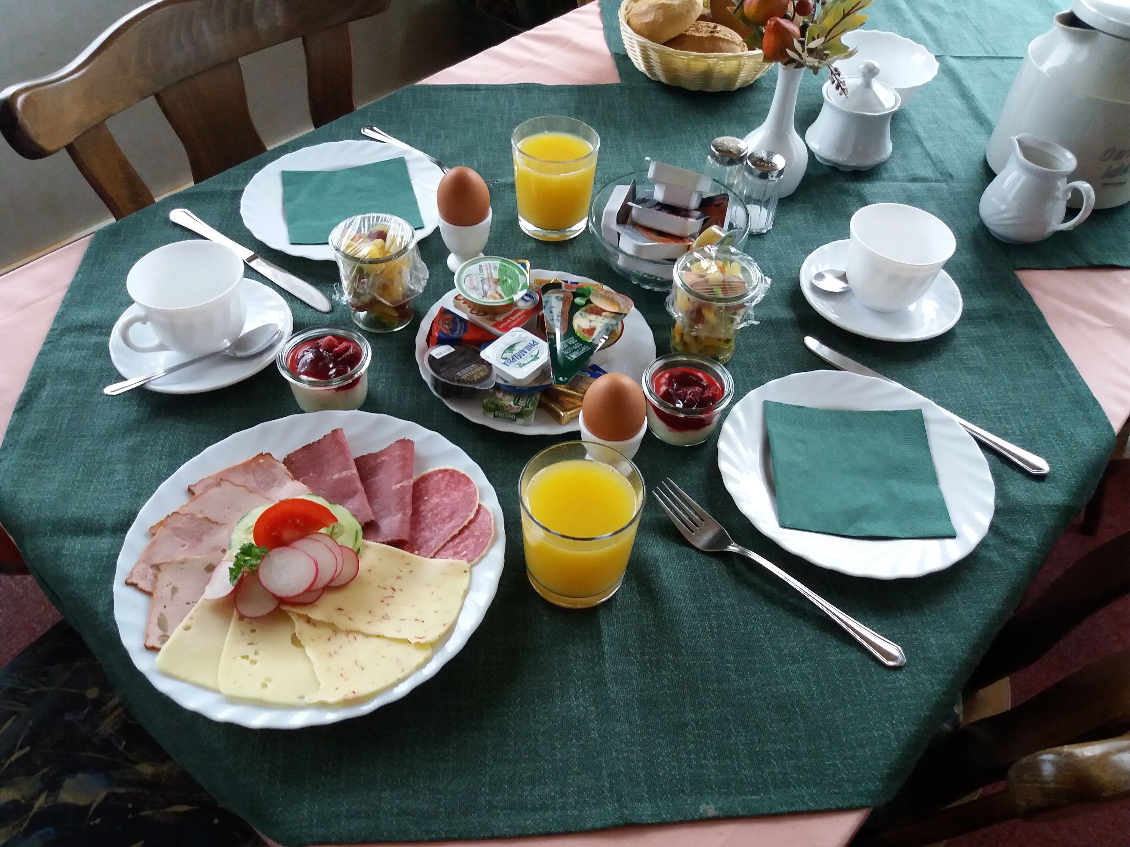 Ein Frühstückstisch mit Tellern voller Speisen, darunter Eier, Schinken, Käse und Obst, sowie Tassen, eine Kanne Orangensaft und diverse Gewürze.