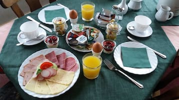 Ein Frühstückstisch mit Tellern voller Speisen, darunter Eier, Schinken, Käse und Obst, sowie Tassen, eine Kanne Orangensaft und diverse Gewürze.
