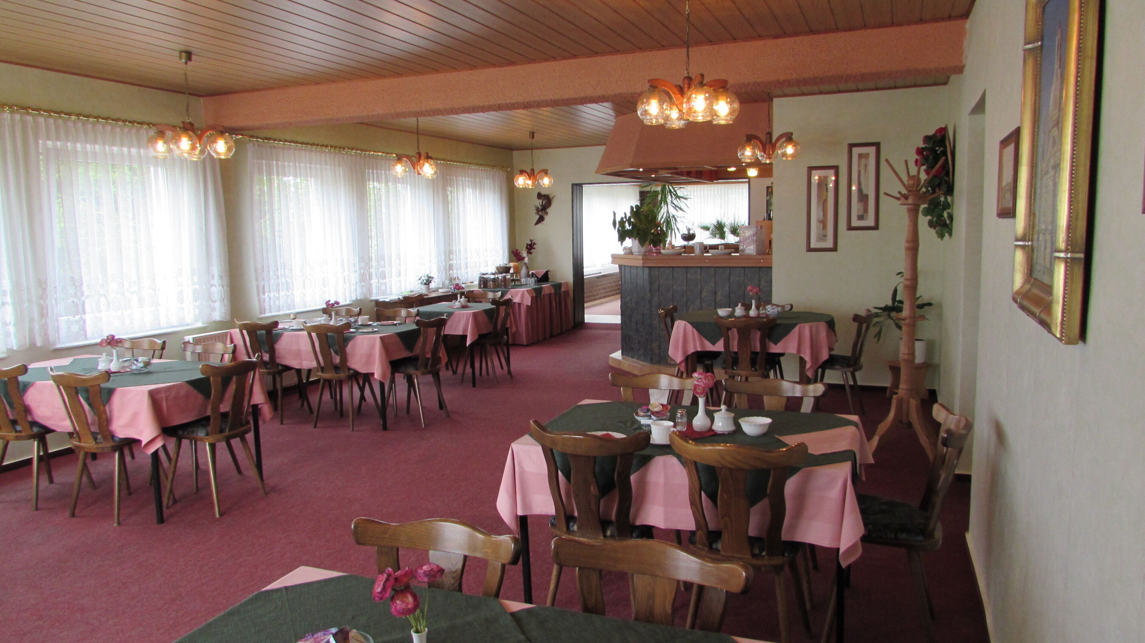 A dining area with multiple tables and chairs, some with pink tablecloths. There are hanging light fixtures and a plant in the center.