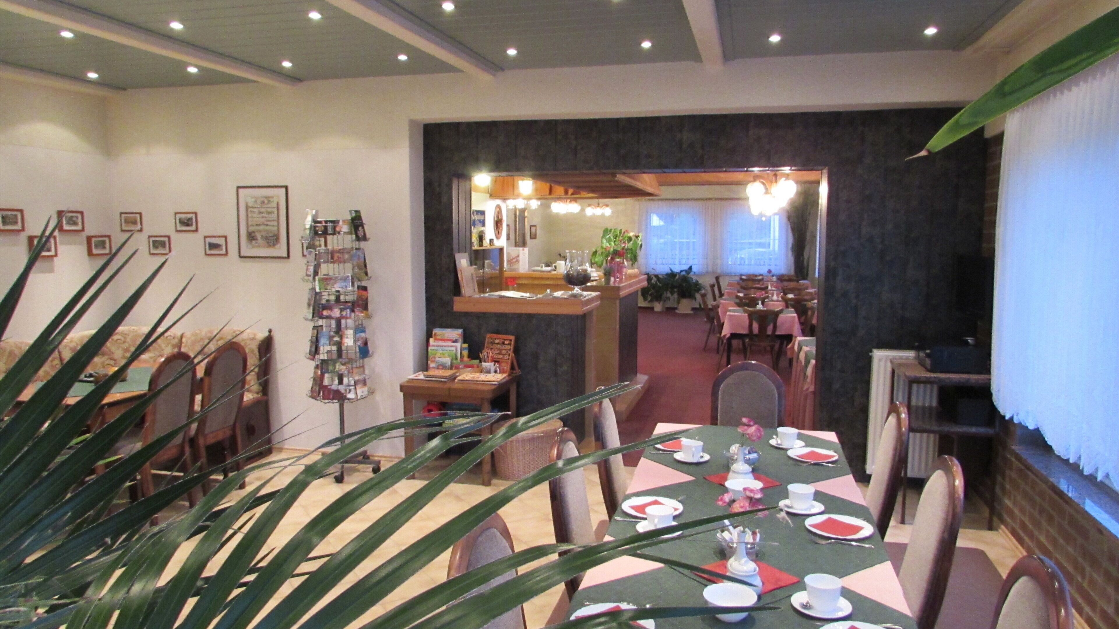 A dining area with tables set for service, a reception desk, framed pictures on the walls, and a plant in the foreground.