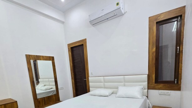 A bedroom with a wooden bed frame, white bedding, a large mirror, a window, and a wall-mounted air conditioner. - Hotel T Star (Meerut)