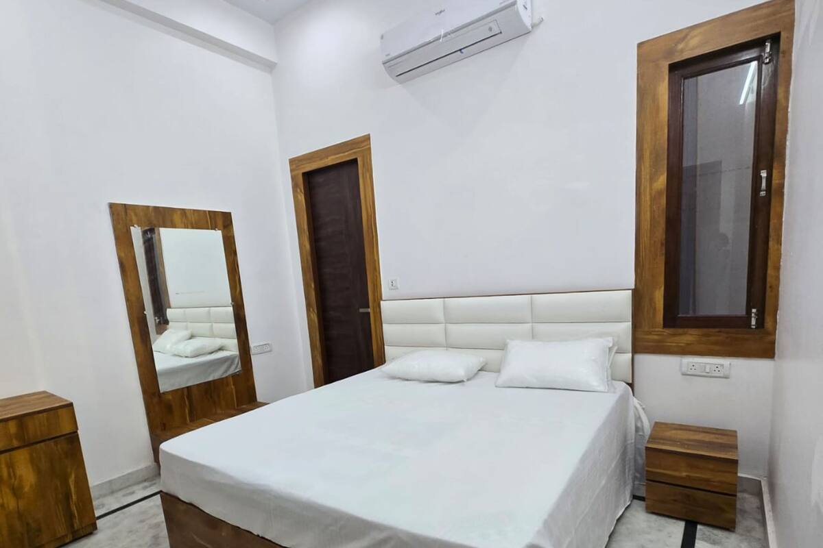 A bedroom with a wooden bed frame, white bedding, a large mirror, a window, and a wall-mounted air conditioner.