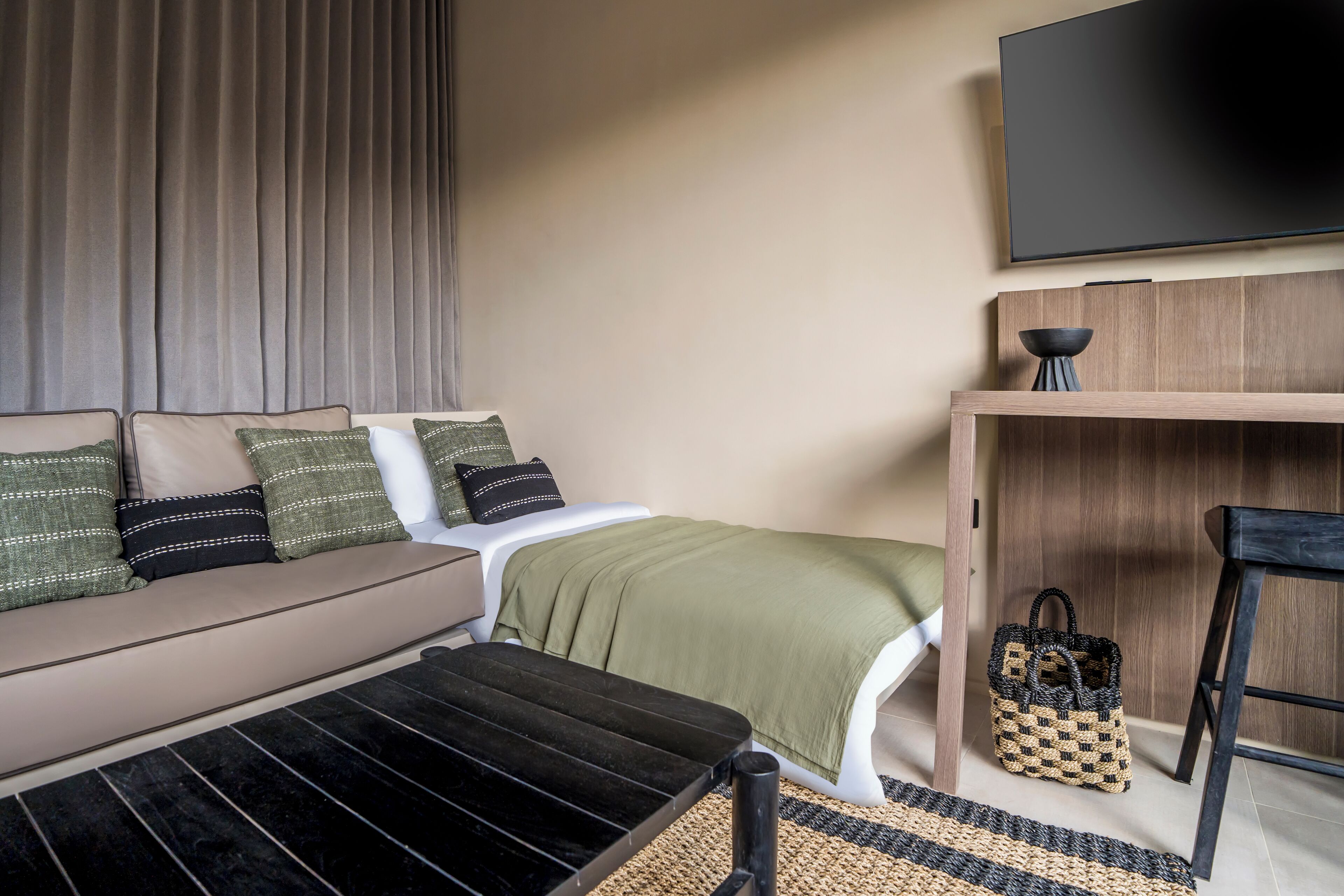 A modern hotel room with a sofa, a small table, a TV, a wooden cabinet, and a woven bag.