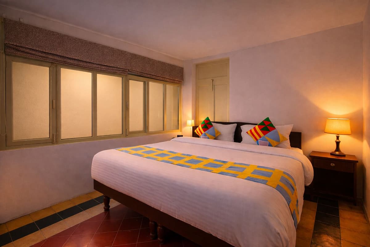 A hotel room with a bed, a nightstand, a lamp, a window with blinds, and a door.