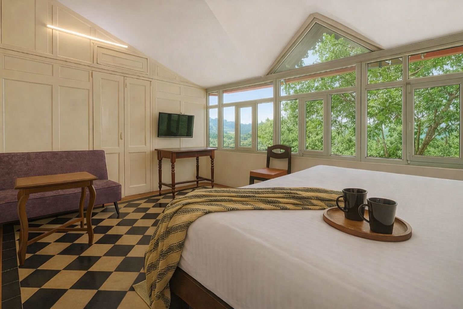 A bedroom with a large bed, a wooden side table, a purple sofa, a TV on the wall, and a window with a view of trees.