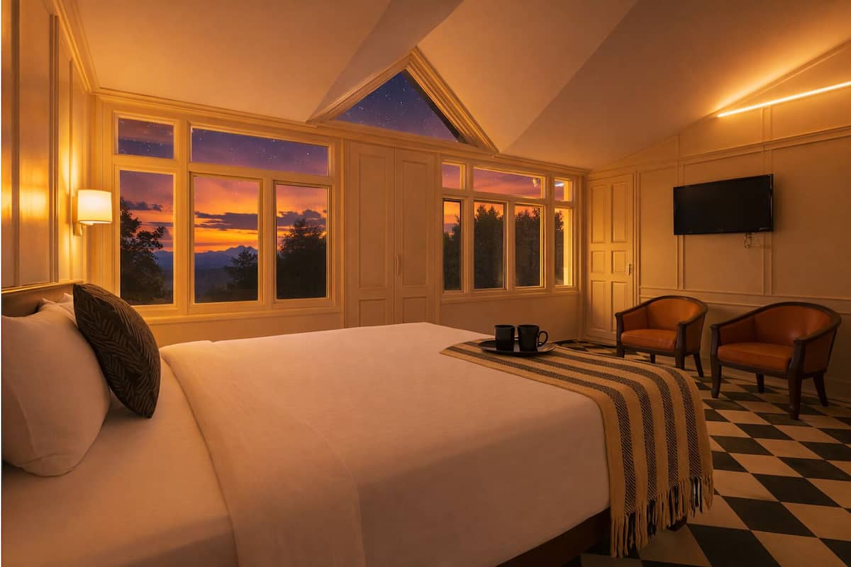 A hotel room with a large bed, two armchairs, a TV, and multiple windows with a view of a sunset.